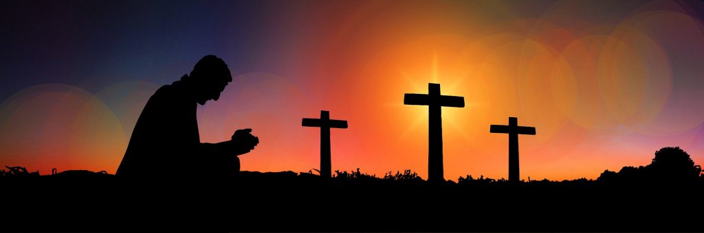 A man kneels in prayer silhouetted in the sunset infront of three crosses on a hill