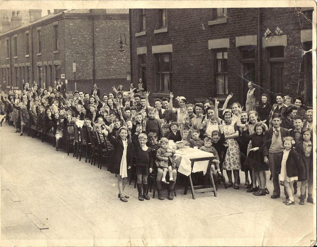 A long line of adults and children face the camera smiling and giving a Victry sign from around a long table down a terraced street.